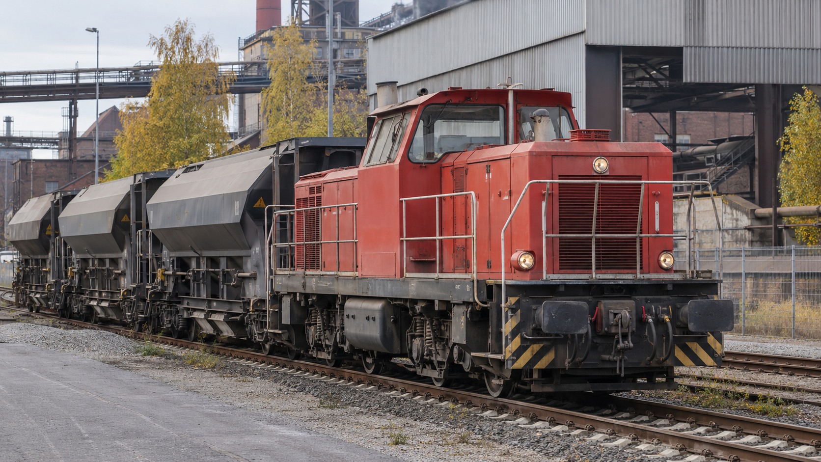 Rote Rangierlok mit Güterwagen vor einer Industriehalle.
